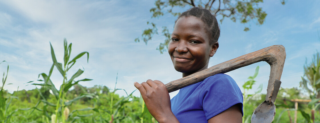 Eine afrikanische Frau hält ein Gartenwerkzeug über die Schulter und steht stolz vor ihrem Maisfeld.