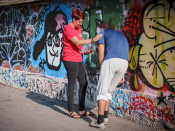 Hilfsperson reicht einer erschöpften Person an einer Graffitiwand Wasser bei großer Hitze und stützt sie im Schatten.