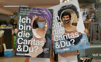 Zwei Mitarbeiterinnen halten zwei große Schiler mit "Ich bin die Caritas & Du" vor dem Gesicht und sehen aus dem ausgeschnittenen Kreis auf diesen Schildern in die Kamera.