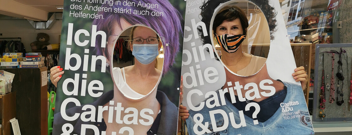 Zwei Mitarbeiterinnen halten zwei große Schiler mit "Ich bin die Caritas & Du" vor dem Gesicht und sehen aus dem ausgeschnittenen Kreis auf diesen Schildern in die Kamera.