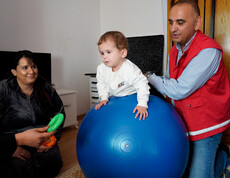 Betreuungsperson stützt ein Kind auf großem blauen Gymnastikball