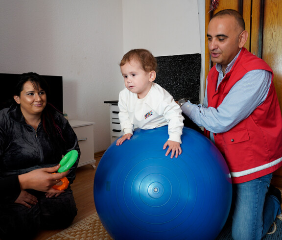 Betreuungsperson stützt ein Kind auf großem blauen Gymnastikball