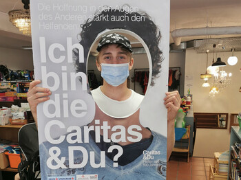 Ein Mitarbeiter sieht durch ein Loch in einer großen Tafel mit dem Schriftzug "Ich bin die Caritas & Du" darauf. 