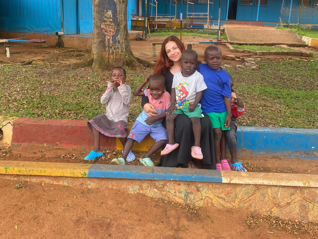 Person sitzt mit mehreren Kindern auf einer bunten Kante im Hof des Nsambya Babies Home.