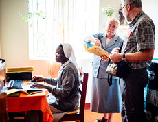 Eine Ordensschwester sitzt vor dem Computer, während eine ältere Ordensschwester dem Diözesanbischof Josef Marketz Unterlagen zeigt.