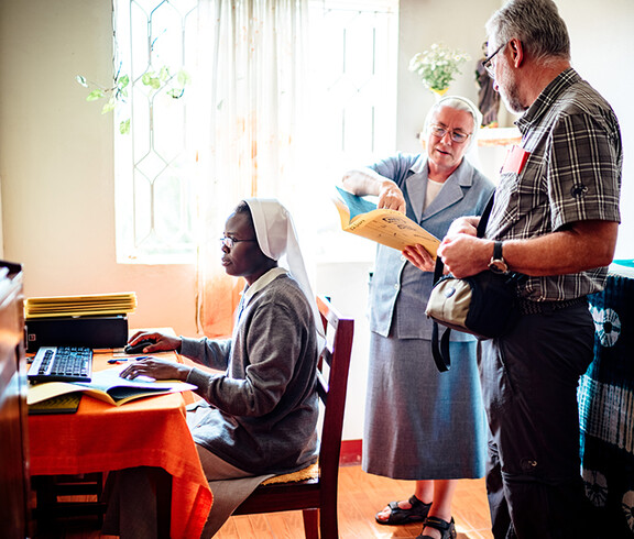 Eine Ordensschwester sitzt vor dem Computer, während eine ältere Ordensschwester dem Diözesanbischof Josef Marketz Unterlagen zeigt.
