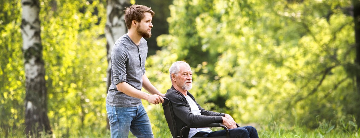 Besuchsdienst bei einem älteren Mann im Rollstuhl  In einem Wald bei schönem Wetter machen ein junger Mann und ein älteren Mann, der im Rollstuhl sitzt, einen Spaziergang.