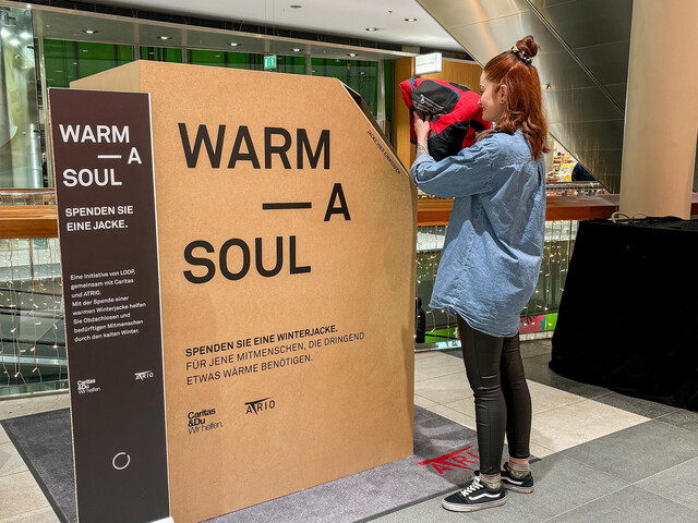 Frau wirft Winterjacke in große Spendenbox mit Text „Warm a Soul“ im Einkaufszentrum ATRIO Villach ein.