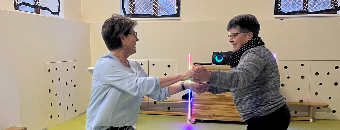 Zwei Damen halten sich an den Händen und tanzen in einem Turnsaal.