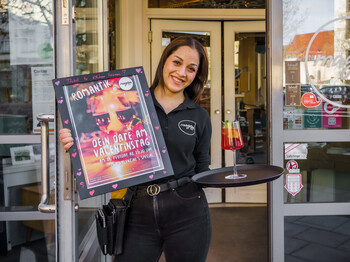 Marwa, Lehrling im magdas LOKAL, hält in der einen Hand das Plakat des Valentinsspecial, in der anderen ein Tablet mit dem Specialdrink.  