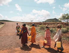 Frauen in bunten Gewändern tragen Wasserkanister auf roter Erde. Im Hintergrund: Hügel, Wolken, weiter Weg.