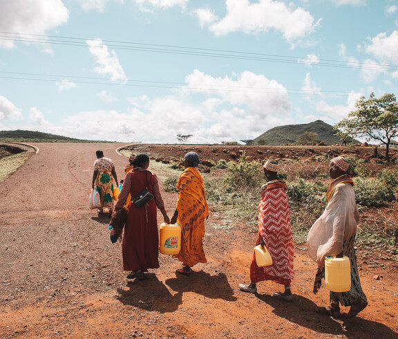 Frauen in bunten Gewändern tragen Wasserkanister auf roter Erde. Im Hintergrund: Hügel, Wolken, weiter Weg.