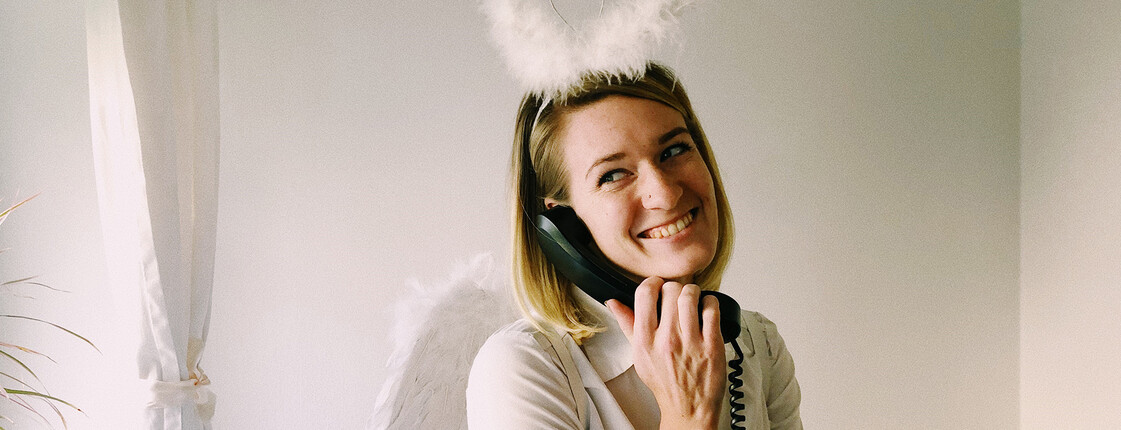 Eine blonde junge Frau mit Heiligenschein und Engelsflügeln telefoniert.