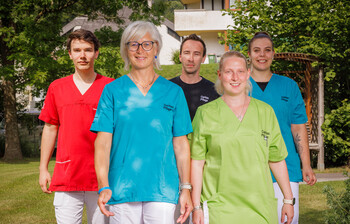 Mitarbeiter*innen in bunten Dienstkleidungen stehen im Garten eines Pflegewohnhauses und lächeln in die Kamera.