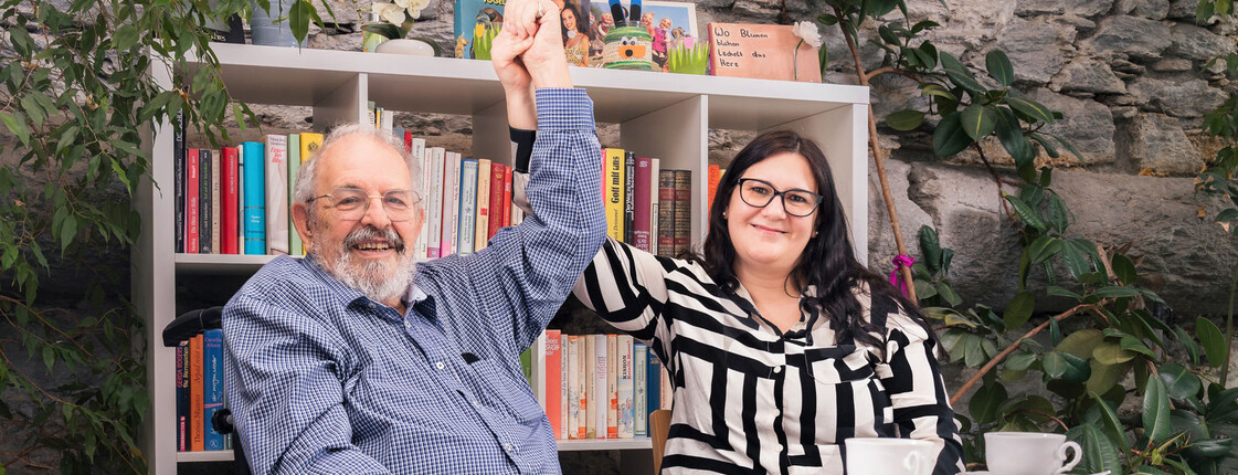 Zwei Personen, eine im Rollstuhl, heben gemeinsam die Hände an einem Tisch mit Kaffee vor Bücherregal zum Thema Pflege und Beruf