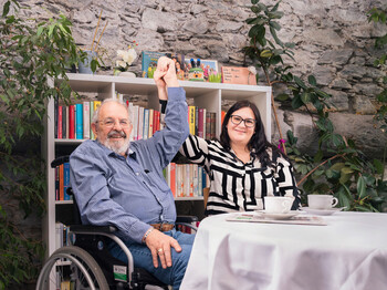 Zwei Personen, eine im Rollstuhl, heben gemeinsam die Hände an einem Tisch mit Kaffee vor Bücherregal zum Thema Pflege und Beruf