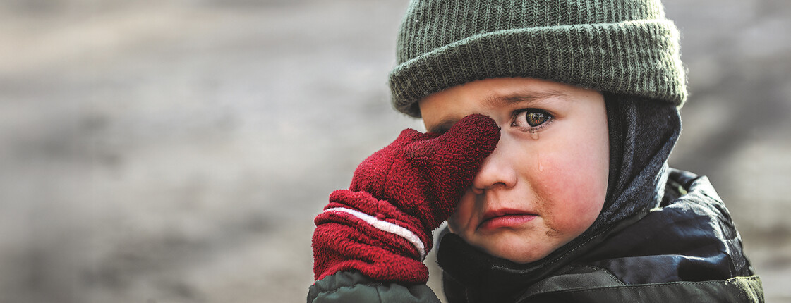 Ein Junge mit einer Schultasche aus Karton steht mitten im Schutt einer Stadt.