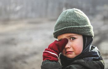 Ein Junge mit einer Schultasche aus Karton steht mitten im Schutt einer Stadt.