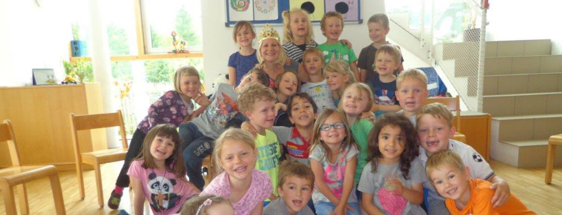 Kinder des Kindergarten Steuerberg sitzen in einem Gruppenraum und lächeln in die Kamera.