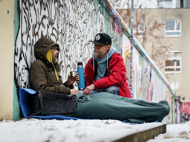 Ein obdachloser Mann sitz im Schlafsack im Schnee an einer Wand, ein Caritas Mitarbeiter reicht diesem ein Heißgetränk.