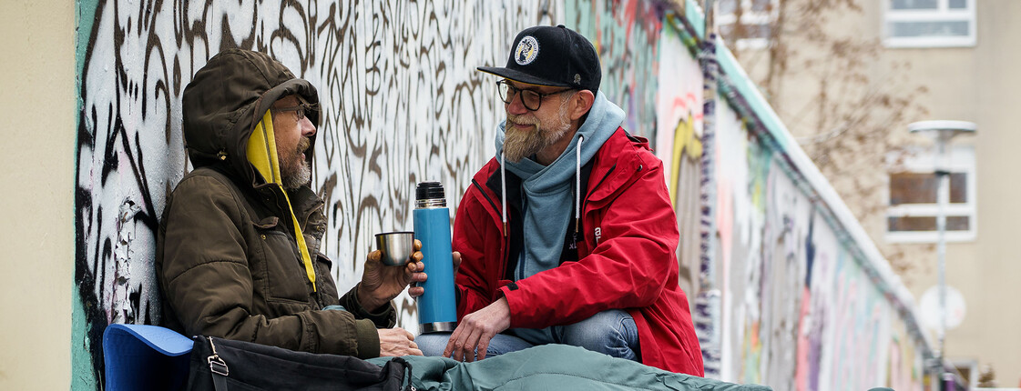 Ein obdachloser Mann sitz im Schlafsack im Schnee an einer Wand, ein Caritas Mitarbeiter reicht diesem ein Heißgetränk.