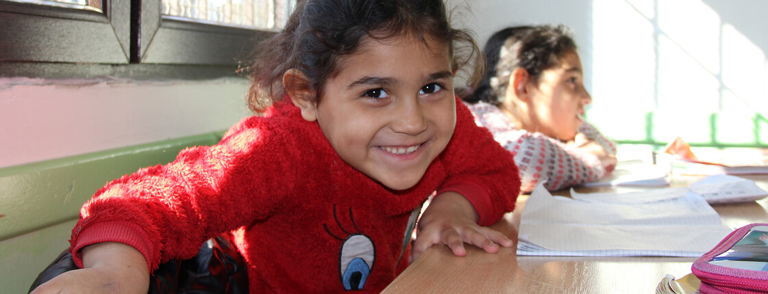 Joscha sitzt an einem Tisch, schreibt konzentriert in ein Heft und hält einen Stift in der Hand. Neben ihr arbeitet ein weiteres Kind an einer Aufgabe. Tageslicht fällt durch ein Fenster auf den Tisch.