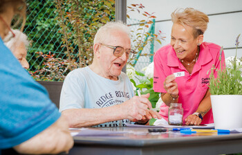 Eine Betreuerin hält einen Zettel mit der Zahl 70 hoch, ein Bewohner kreuzt auf seinem Bingo-Zettel diese Zahl ab.
