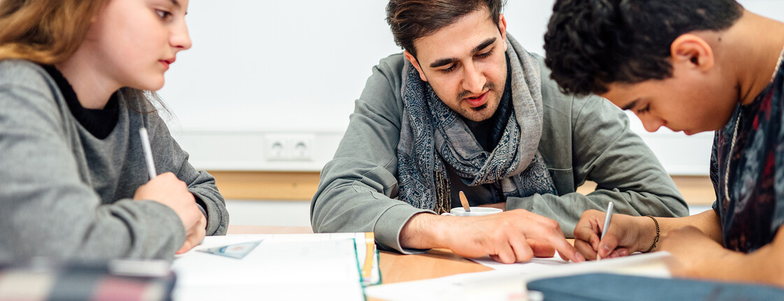 Ein Lernhelfer sitzt mit zwei Lerncafé Schüler*innen am Tisch und hilft ihnen bei den Hausaufgaben.