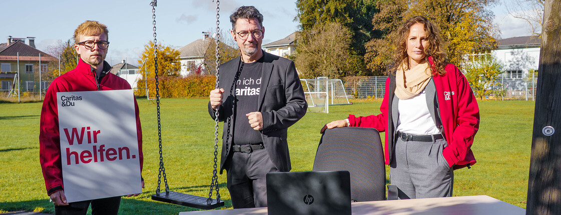 Drei Caritas-Mitarbeitende mit Tisch und Plakat am Spielplatz als Zeichen gegen Care-Arbeits-Überlastung.