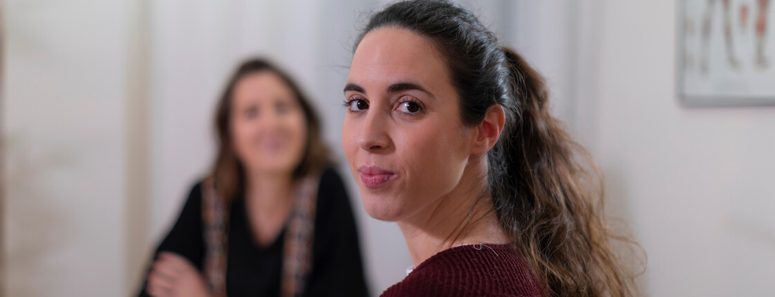 Portrait einer Frau mit dunklen Haaren, die über ihre Schulter in die Kamera schaut während sie sich in einem Beratungsgespräch befindet.