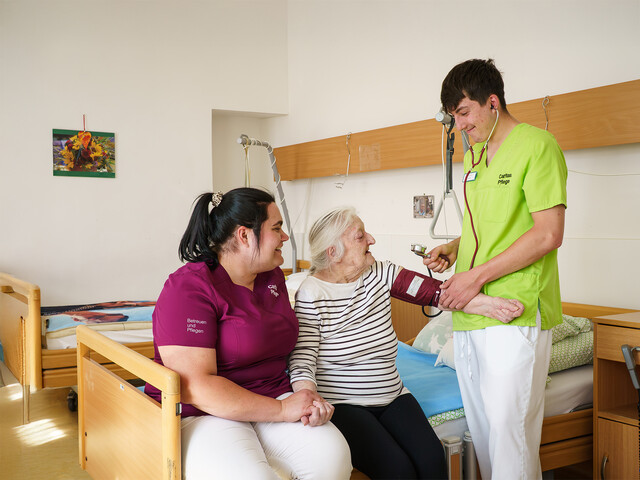 Zwei Pflegekräfte begleiten eine ältere Person beim Blutdruckmessen in einem Pflegezimmer der Caritas.