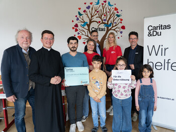 Kleines Fest zur Eröffnung mit Hausherrn Christian Reiter, Stadtpfarrer Christoph Kranicki, Lernhelfer Human, Lerncafé-Leiterin Dörte Enzi, Mondi Frantschach-Geschäftsführer Tobias Sollanek, Lerncafé-Mitarbeiterin Ines Vormaier, Caritasdirektor Ernst Sandriesser und Lerncafékindern (v.li). Gruppe aus Kindern, Freiwilligen und Unterstützer*innen im Lerncafé Mondi Wolfsberg vor Caritas-Rollup und Wandbild mit Herzen.