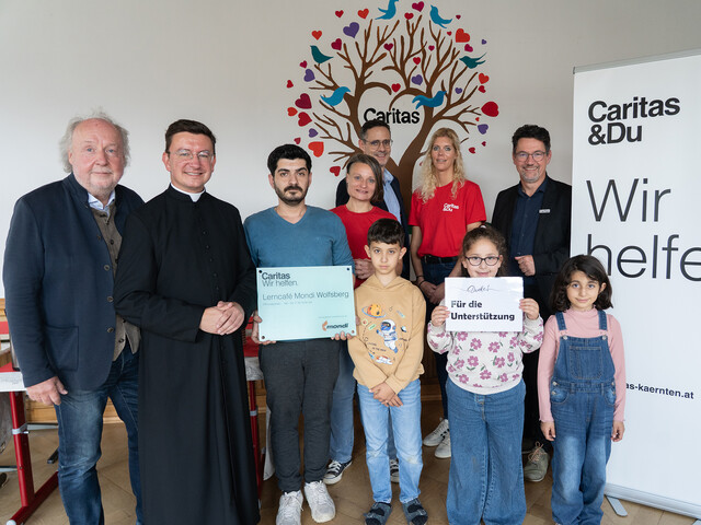 Gruppe aus Kindern, Freiwilligen und Unterstützer*innen im Lerncafé Mondi Wolfsberg vor Caritas-Rollup und Wandbild mit Herzen.