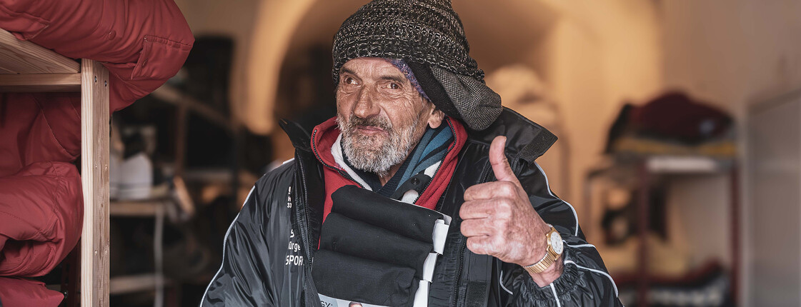 Ein obdachloser Mann probiert eine neue Jacke an und zeigt einen Daumen nach oben, während er lächelt.