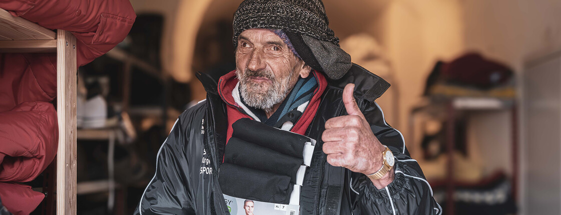 Ein obdachloser Mann probiert eine neue Jacke an und zeigt einen Daumen nach oben, während er lächelt.