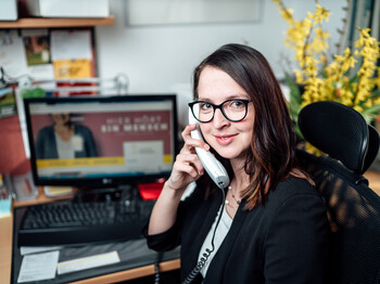 Eine junge Frau sitzt vor einem Computer und hält einen Telefonhörer in der Hand.