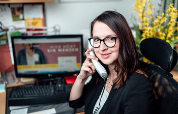 Eine junge Frau sitzt vor einem Computer und hält einen Telefonhörer in der Hand.
