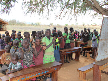 Schüler*innen sitzen zwar im Freien, aber auf den neu angeschafften Schulbänken vor der Tafel und klatschen im Rythmus.
