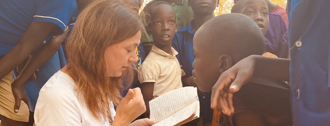 Caritas-Auslandshilfe-Bereichsleiterin Alexandra Blattnig-Rull spricht mit Kindern in Uganda und notiert etwas in ihrem Notizbuch.