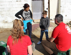 Caritas-Projektreferentin Theresa Sacher steht mit einem Jungen und einer Frau vor einem einfachen Haus. Zwei weitere Erwachsene sitzen oder stehen gegenüber, während ein Gespräch stattfindet.