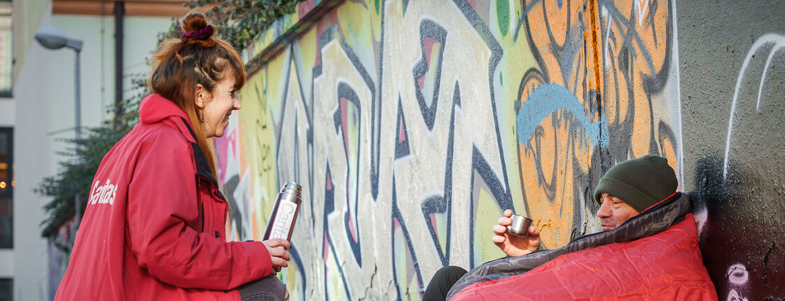 Eine Caritas-Mitarbeiterin bringt einem obdachlosen Mann warmen Tee – Hilfe und Menschlichkeit direkt auf der Straße.