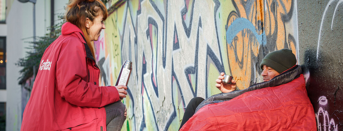 Eine Caritas-Mitarbeiterin bringt einem obdachlosen Mann warmen Tee – Hilfe und Menschlichkeit direkt auf der Straße.