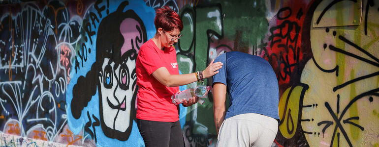 Hilfsperson reicht einer erschöpften Person an einer Graffitiwand Wasser bei großer Hitze und stützt sie im Schatten.