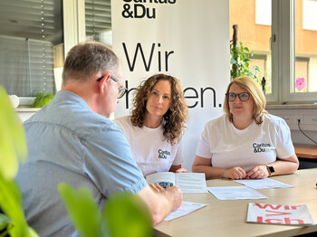 Zwei Mitarbeiterinnen sitzen mit einem Mann, der sein Hab und Gut bei einem Hochwasser verloren hat, an einem Tisch und füllen gemeinsam Formulare für die Hilfsleistungen aus.