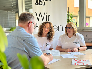Zwei Mitarbeiterinnen sitzen mit einem Mann, der sein Hab und Gut bei einem Hochwasser verloren hat, an einem Tisch und füllen gemeinsam Formulare für die Hilfsleistungen aus.