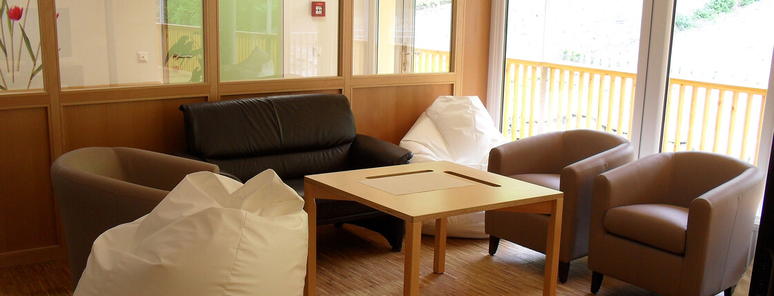 Ein Gemeinschaftsraum in dem ein Tisch in der Mitte steht, dahinter ein Sofa und rechts zwei Sessel. Auf der rechten Seite steht ein Sitzsack. Alles steht vor der Tür zum Balkon in den schönen Garten.