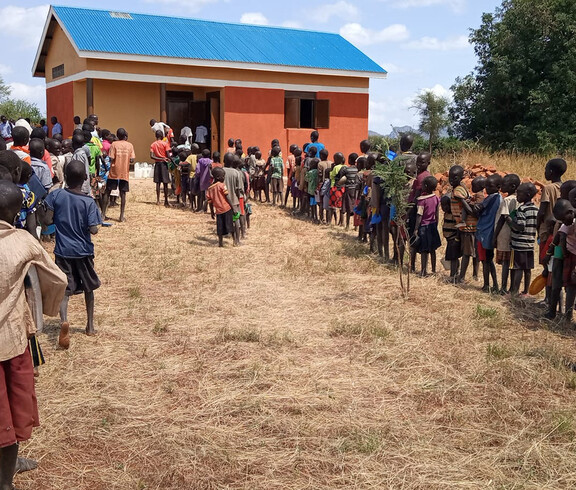 Viele Schulkinder stehen in einer langen Reihe vor einem kleinen Gebäude mit blauem Dach und warten auf ihre Mahlzeit.