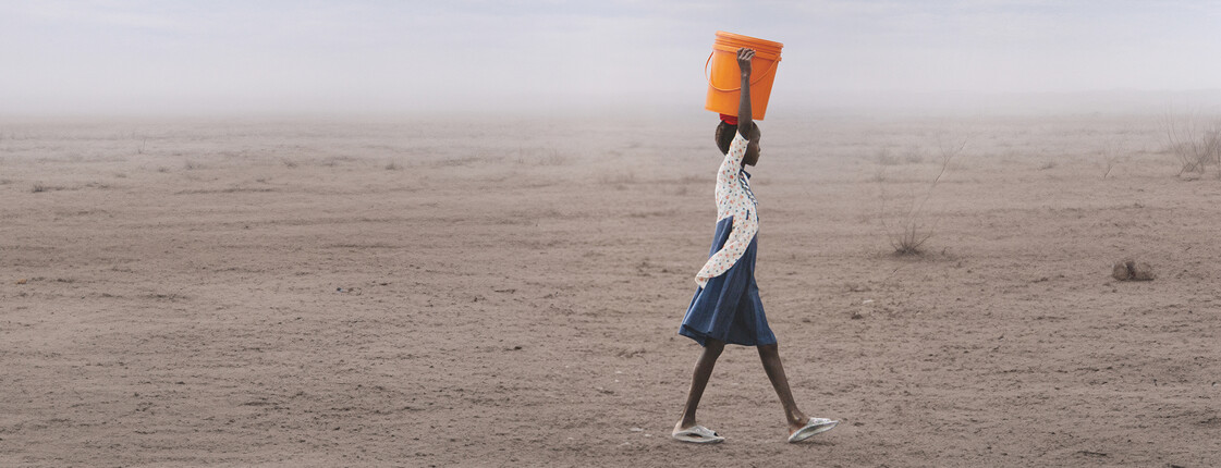 Person trägt orangefarbenen Wasserkanister durch ausgetrocknete Landschaft unter weitem Himmel, Thema Hunger und Wasserknappheit.
