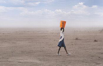 Person trägt orangefarbenen Wasserkanister durch ausgetrocknete Landschaft unter weitem Himmel, Thema Hunger und Wasserknappheit.
