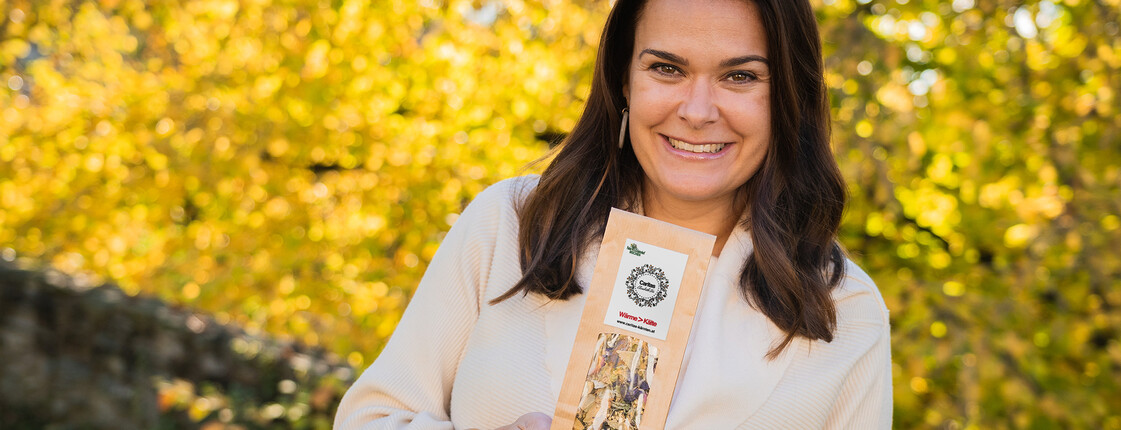 Eine junge Frau hält den Caritas-Kräutertee in der Han dund steht vor herbstlichen Bäumen.